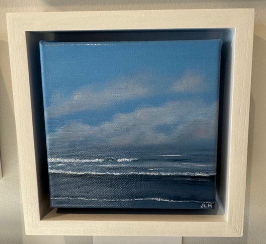 A square canvas painting of a blue ocean and cloudy sky, mounted in a white shadow box frame on a beige wall.