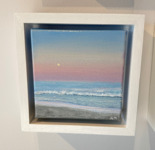 A square framed beach sunset painting mounted on a light gray wall. The artwork shows soft pastel waves, a pale blue sky, pink and peach horizon, and a small sun, set in a white float frame.